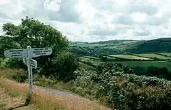 White signpost on a path on the left. To the right are rolling green fields on the hillsides.