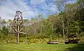 A wooden derrick at left on grass, with trees and a mountain ridge in the background