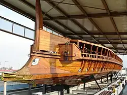 A galley sitting on struts on land under in an open-sided shed seen from the front; it has a large cast bronze ram along the waterline and an eye of Horus painted above the ram.
