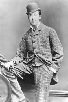 Oscar Wilde posing for a photograph, looking at the camera. He is wearing a checked suit and a bowler hat. His right foot is resting on a knee-high bench and his right hand, holding gloves, is on it. The left hand is in the pocket.