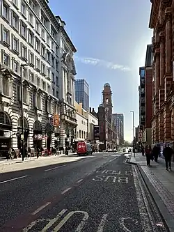 A photograph of a wide road from the right-hand pavement. Tall buildings tower on either side, with a white stucco building on the left and a red brick building on the right. A church tower is visible in the background in front of a church building.