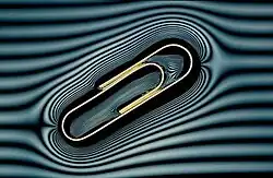 A metal paperclip floating on water. A grille in front of the light has created the 'contour lines' which show the deformation in the water surface caused by the metal paper clip.