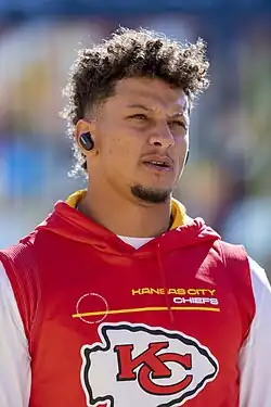 Patrick Mahomes standing on a football field. He is wearing a white jersey with a red number "15" and red sleeves.