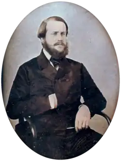 Photographic half-length portrait of a seated bearded man dressed in a dark, double-breasted coat with his right hand tucked inside the front