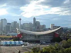 Exterior shot of an indoor arena. The building has a sloped roof in the shape of a reverse hyperbolic paraboloid and a primarily-concrete outer facing with red towers at the corners. Several skyscrapers are visible in the background.