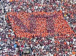 People in the stadium wearing orange and maroon T-shirts to form an image of the letters "VT" in maroon on a square background of orange.