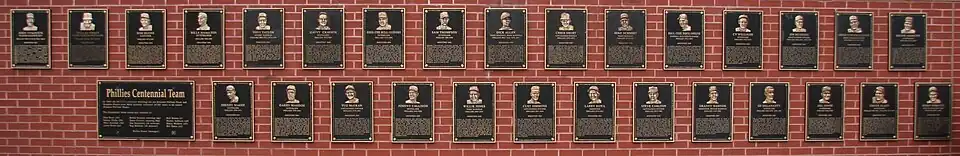 Forty black metal plaques are mounted on a brick wall. They are inscribed with gold images of human faces and text.