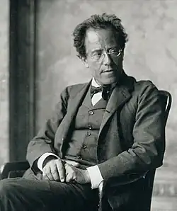 Middle-aged man, seated, facing towards the left but head turned towards the right. He has a high forehead, rimless glasses, and is wearing a dark, crumpled suit