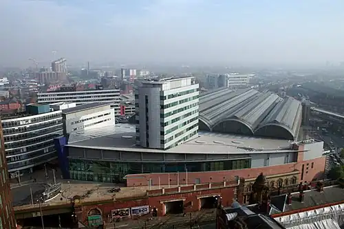 An aerial photo of a modern railway terminus station viewed from the top end a short distance away. The multiple parallel roofs of the trainshed stretch into the background against a grey sky.