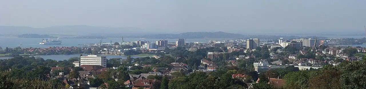 A panorama of Poole town centre viewed from Parkstone, showing some tall buildings and the rooftops of low-rise buildings amongst treetops, with a ship in the harbour beyond.