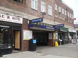 A brown-bricked building with a rectangular, dark blue sign reading "QUEENSBURY STATION" in white letters all under a light blue sky
