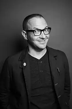 Black-and-white portrait of Cory Doctorow, a white man in his late 40s. He smiles at the camera with raised eyebrows, wearing dark-rimmed glasses, a suit jacket with a button on the lapel and a pen in the breast pocket over a button-up T-shirt. A headset microphone is visible, and his hair is cut short in a buzz cut.