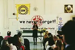 A seated crowd facing a standing woman. Behind her is a table with flowers. Above the table is a large banner with the text, "We never forget!" along with the IWW name and globe logo. A variety of United Auto Workers logos are visible on the wall in the background.