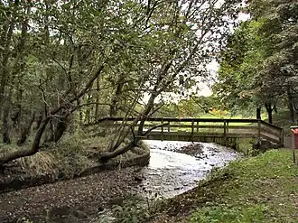 View down a small river, bending to the left, crossed by a wooden footbridge in the middle distance.
