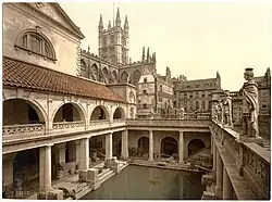 A late-nineteenth-century Photochrom of the Great Bath at the Roman Baths. Pillars tower over the water, and the spires of Bath Abbey&nbsp;– restored in the early sixteenth century&nbsp;– are visible in the background.
