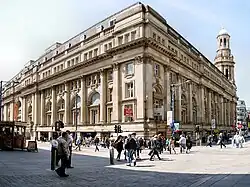 A palladian square building as viewed from its corner. Outside the front of the building is a large pedestrianised square which is quite busy with people. The back right corner of the building continues above the rest of the flat roof to become a conical tower.
