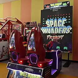 Two red chairs mounted on a base face a large screen displaying "Space Invaders Frenzy". Plastic toy cannons with lights are placed in front of the chairs aimed at the screen.