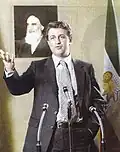 Sadeq Tabatabaei speaking at a post-revolution press conference with the Lion and Sun flag behind him