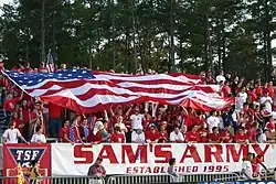 U.S. soccer fans, dressed in red, cheer in bleachers as they hold a large U.S. flag over themselves at a soccer match.