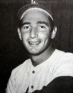 Black and white photo of a man in a white baseball jersey and dark baseball cap with an interlocked white "LA" on the front, smiling at the camera.