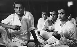 Photograph of Satyajit Ray seated with Ravi Shankar with several others in the background