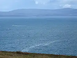 The attack site today, seen from a cliff above the bay. A small green wreck buoy is a few hundred metres away. A thin slick of oil is on the surface of the sea.