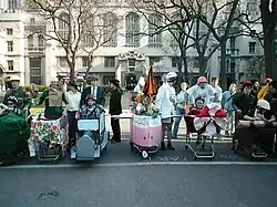 A group of students in front of six shopping carts that have been decorated; there is a student wearing a helmet in each one
