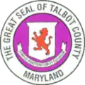 A round seal featuring a long on a shield and ribbon against a purple background with a white border