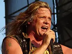 A middle-aged Caucasian man with long, blond hair and tattoos on his right shoulder wearing a sleeveless khaki shirt sings into a microphone.