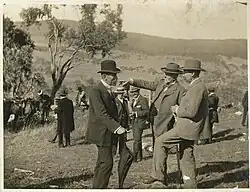 Three middle-aged men with short beards in formal suits and hats standing in open hilly field with single tree nearby
