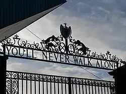 Design of the top of a set of gates, with the sky visible. The inscription on the gates reads "You'll Never Walk Alone".
