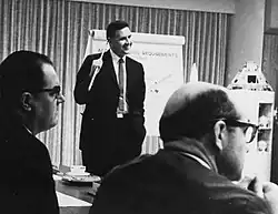 A cropped black and white photograph of a standing suited man explaining something to at least two seated suited men