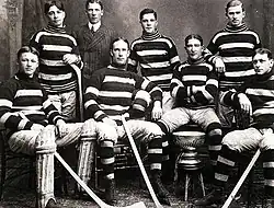 Seven men in hockey uniforms, four sitting in the front row, around the Stanley Cup trophy. Three stand in the back, with a man in a suit standing behind them.