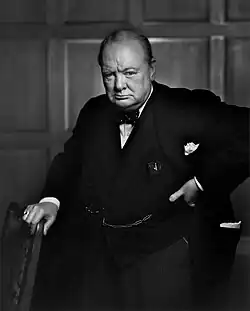 Three-quarter length portrait photograph of Churchill, 67, wearing a suit, standing and holding onto the back of a chair