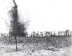 In the monochrome photograph, a shell detonates amidst a field strewn with multiple layers of tangled strands of barbed wire.