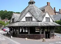 A small single-story building with a pyramid shaped roof, to the side of a road lined with buildings. Some private small cars are visible. Trees in the distance with the skyline of Dunster Castle.
