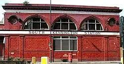A two-storey red-glazed building with a single rectangular window on the lower floor and three large semi-circular windows on the upper floor flanked by two small circular windows. A white band between the floor levels displays "South Kensington station".
