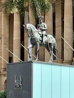 Equestrian statue in dark grey metal of George&nbsp;V in military dress uniform on a plinth of red granite outside a Classical building of red sandstone