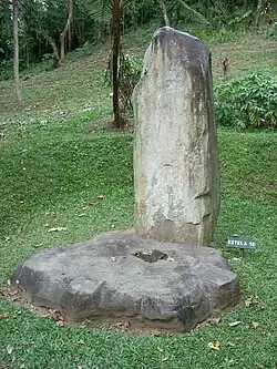 A tall, narrow standing stone set in a grassy area with an overgrown mound in the background. A flat circular stone lays at the base of the standing stone, it has been carved into a rough cogwheel shape.