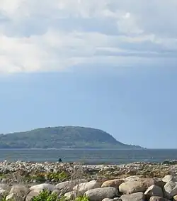 A grey-blue body of water separating smooth, large, light-coloured rocks in the foreground and a hazy green hill in the background, all under a blue sky with white clouds.