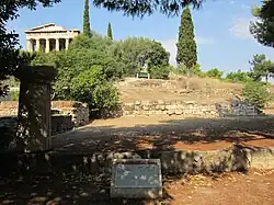 Color photograph of a ruin in the open. A single Greek column can be seen in the foreground on top of a compound wall. In the background are some trees, and at the rear left is a Classical Greek building.