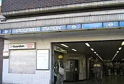 A brown-bricked building with a rectangular, light blue sign reading "STOCKWELL STATION" in white letters all under a light blue sky