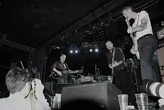 Swans performing live in Warsaw in December 2010. From left to right: Kristof Hahn on lap steel, Michael Gira on guitar, Thor Harris on percussion, Chris Pravdica on bass guitar, and Norman Westberg on guitar. Phil Puleo is seated behind the drum kit.