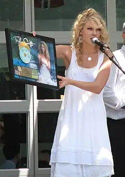 Taylor Swift speaking into a mic, wearing a white sundress while holding her album