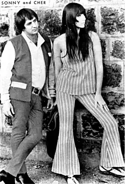 Sonny and Cher stand against a stone wall; Sonny wears a vest over a white shirt and dark pants, while Cher wears a striped sleeveless top with matching flared pants.