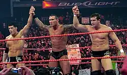 Three dark-haired men stand side-by-side in a wrestling ring with red ropes. The man in the center has tattoos on both his arms which are being raised in the air by the two men on either side of him. All three are wearing black wrestling tights.