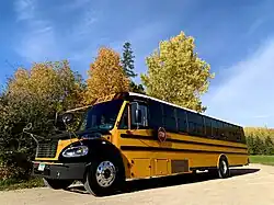 A distinctive yellow-orange bus is parked on the edge of a country road in front of a background of leafy trees.