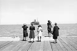 Five women standing at the edge of a dock. A large ship is sailing away from them.