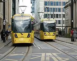 Two yellow trams sit besides one another on parallel tracks, with the one on the left slightly closer to the camera which is on the ground in the road. Both have their pantographs up which are touching contact wires. On either side there are people walking on the pavement.