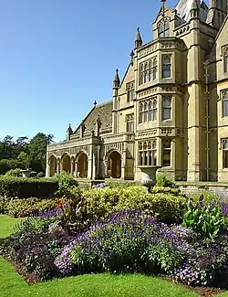 Yellow stone ornate facade of building with lower arched front to the left. In the foreground could be flowers in a formal garden.